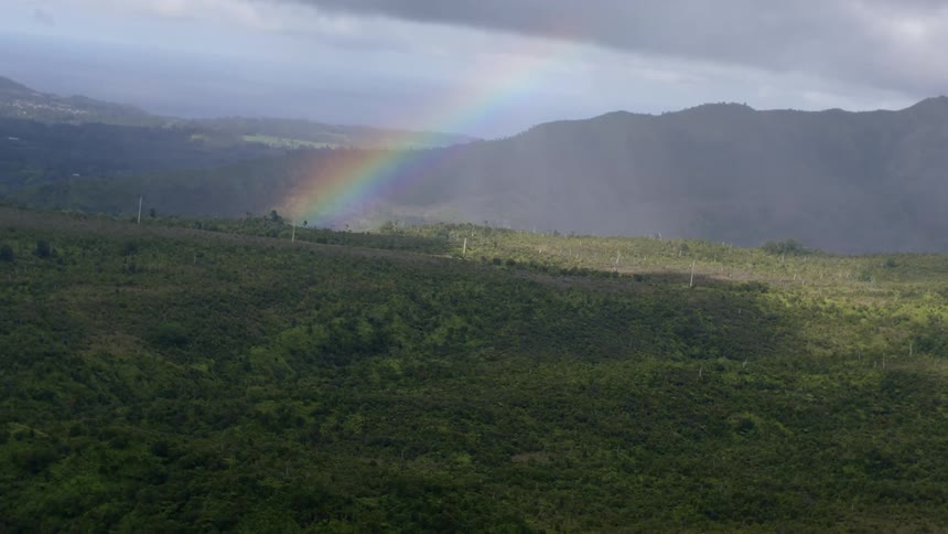 An aerial view shows a vibrant rainbow arcing across a landscape of green hills and mountains under a stormy sky