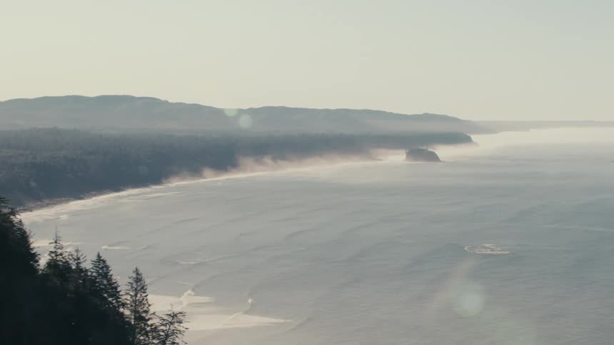 A hazy panoramic view of a coastline stretches into the distance with a long sandy beach bordering a calm ocean bay fading into a misty horizon under a pale sky