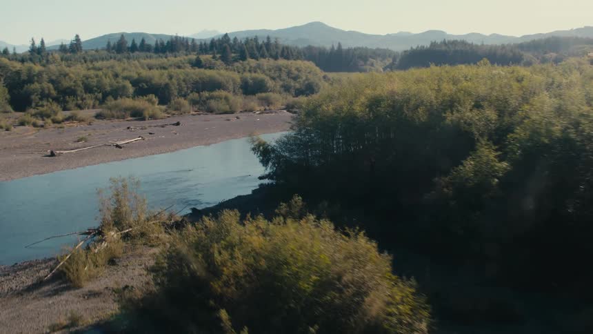 An aerial view shows a winding river course through a barren landscape revealing exposed earth and scattered debris alongside patches of remaining forest