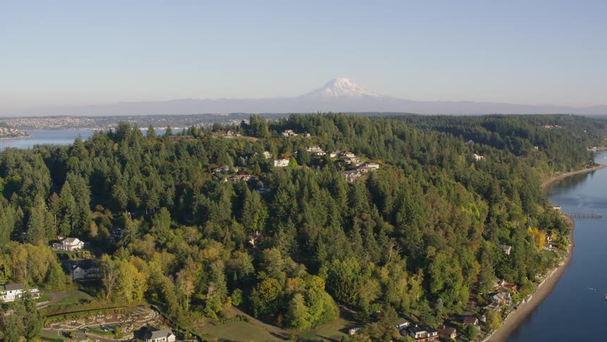 A distant snow capped mountain peak overlooks a densely forested island in a calm lake with a sprawling cityscape visible in the hazy distance under a clear sky