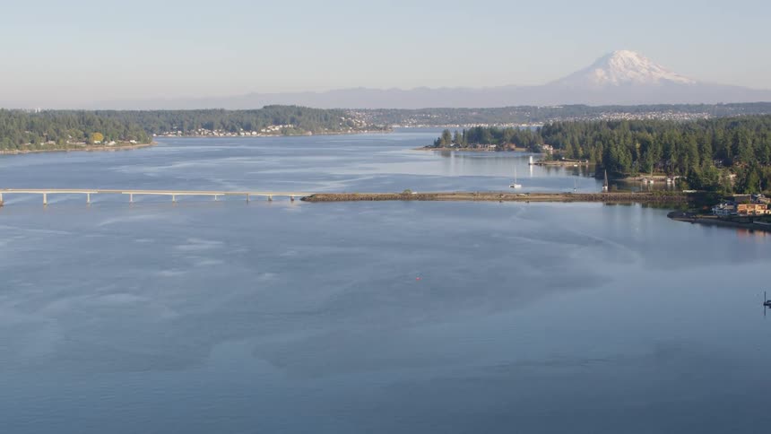 An aerial view shows a wide lake with a bridge spanning it a distant snowcapped mountain and surrounding shoreline