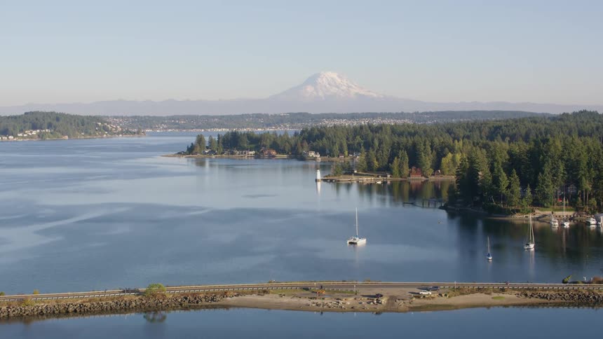 A majestic snow capped mountain stands sentinel in the distance overlooking a serene lake dotted with sailboats and surrounded by lush forests and a causeway