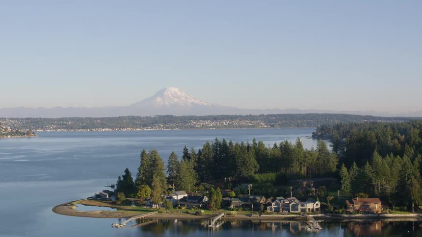 A distant snow capped mountain looms over a tranquil lake dotted with green islands and surrounded by a sprawling city all under a clear blue sky