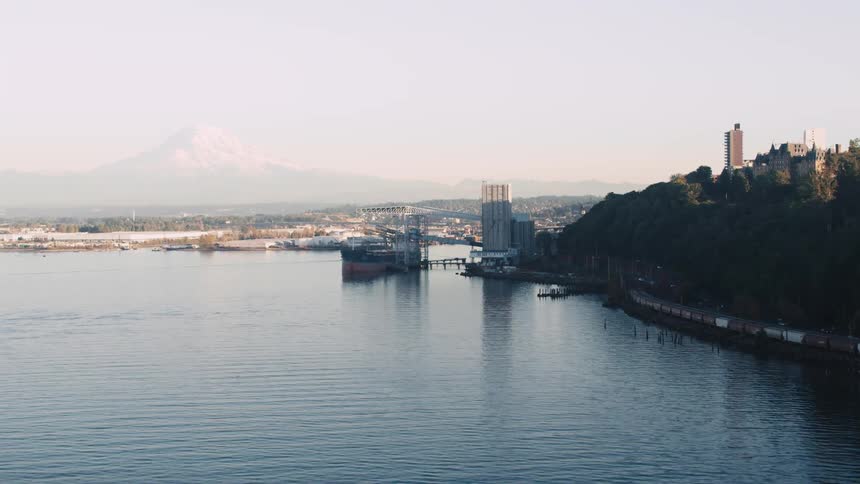 Two grey supply ships docked in a harbor a city on a hill to the right a snow capped mountain in the distant background
