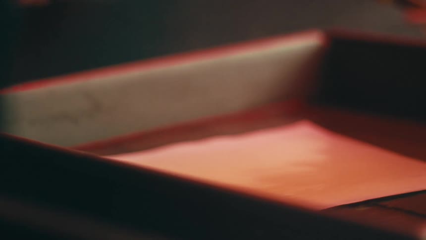 A person wearing an apron handling a piece of paper or film in a tray filled with liquid as part of a photographic development process