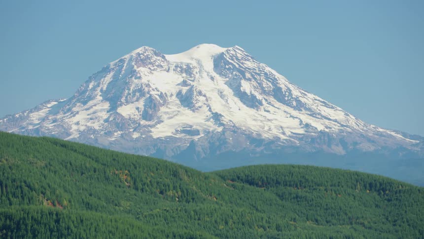 A large snow capped mountain behind a ridge covered in lush green forest