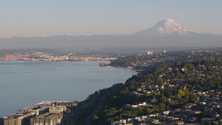 An elevated view captures a sprawling cityscape bordering a calm bay with a majestic snowcapped mountain dominating the distant horizon