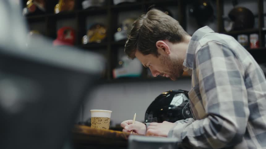 A young man in a plaid shirt deeply focused as he writes on paper at a wooden counter with a black helmet beside him and a coffee cup nearby while shelves filled with various helmets line the background