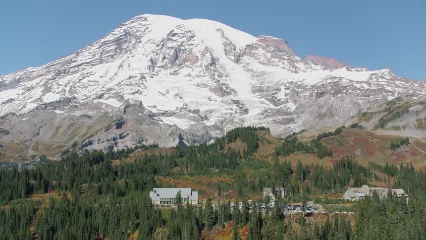 A snow covered mountain with evergreen trees and autumn colors at the base and three buildings with a parking lot full of cars