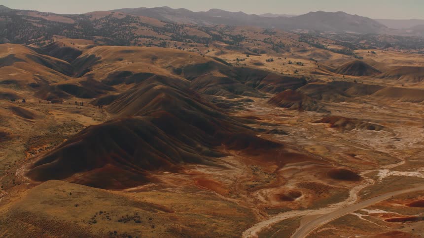 A vast undulating landscape of barren hills and sparse vegetation stretches to the horizon under a muted sky with a small winding path visible
