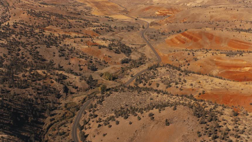 A winding road snakes through a barren hilly landscape dotted with sparse vegetation and patches of reddish soil under a vast sky