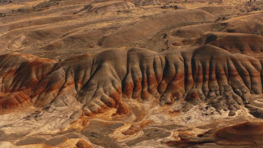A rugged arid landscape with rolling hills and exposed rock formations stretches under a mostly clear sky displaying a blend of earthy tones and textures