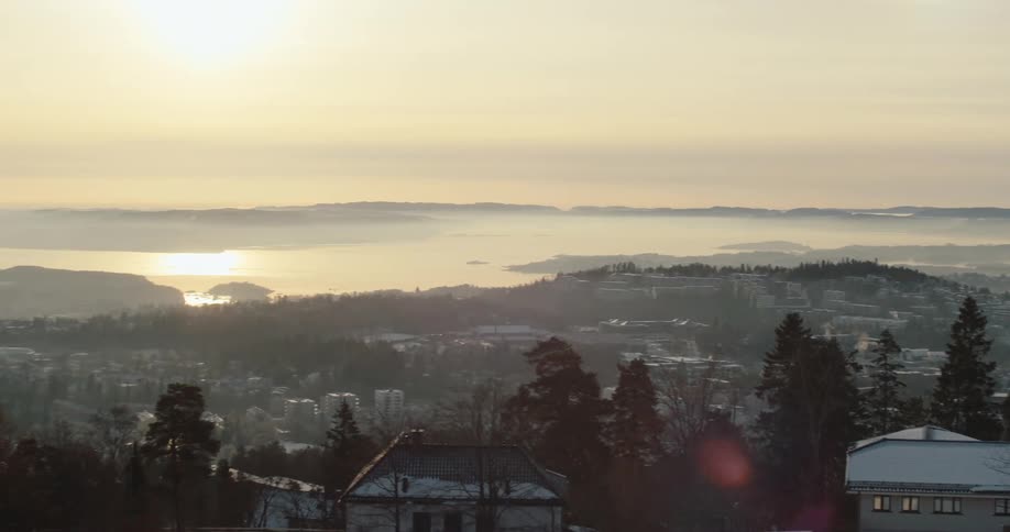 A panoramic view of a city nestled among hills with snowcovered rooftops and trees in the foreground and a layer of fog or mist in the distance