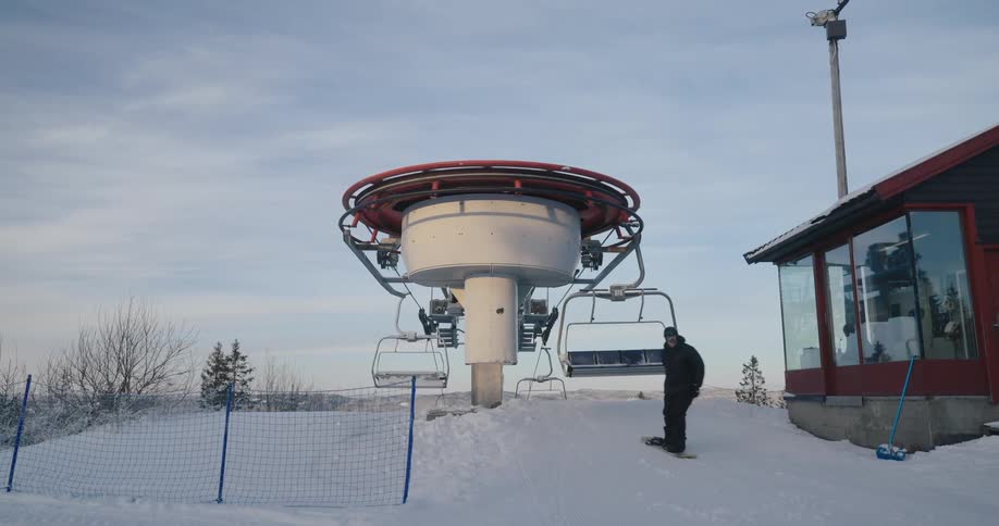 A person steps off a ski lift onto a snowy slope with the lifts machinery and a small building in the background