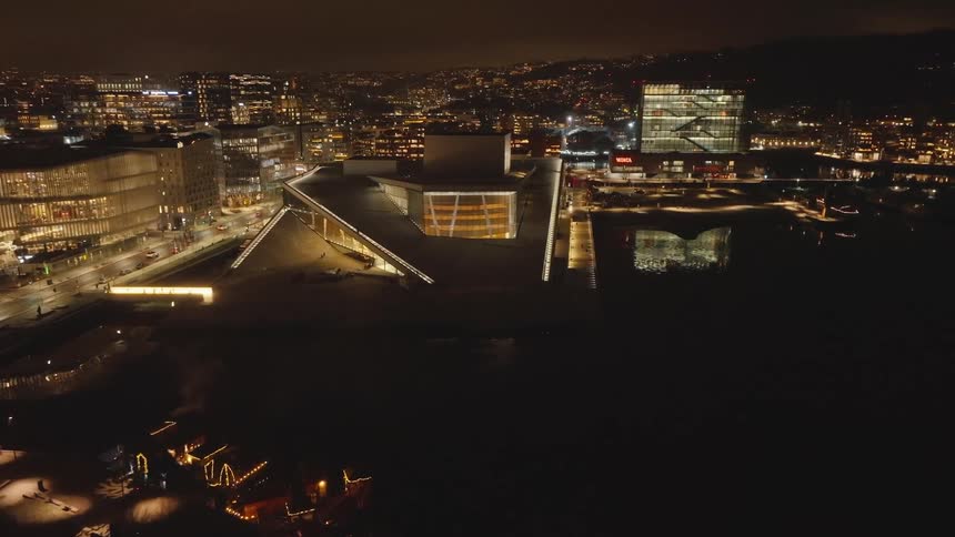 Large building with angular architecture sits on the waterfront near a highway in a city at night