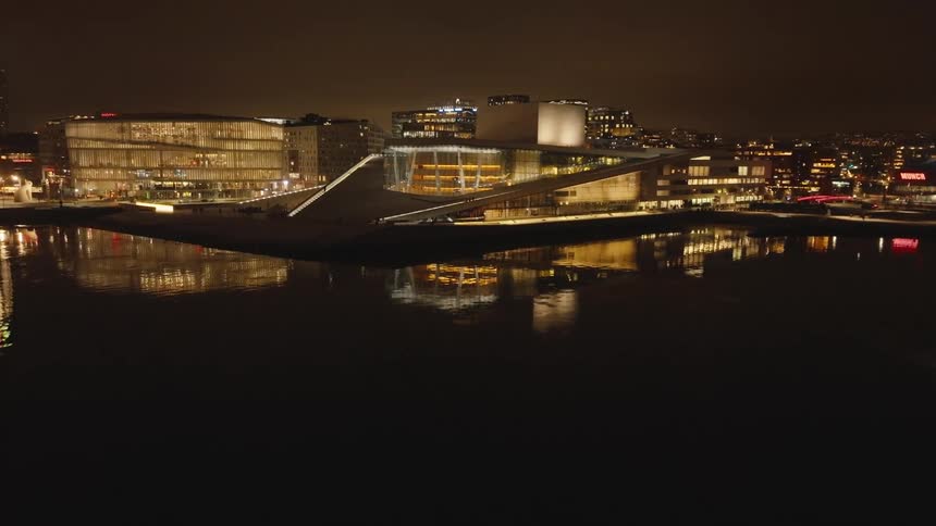 The oslo opera house with its distinctive angled white marble exterior and illuminated interior is captured at night reflected in the calm waters of the surrounding fjord with the city lights of oslo