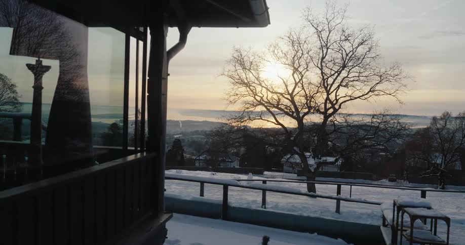A serene winter landscape with a snowcovered yard a bare tree and a distant view of a lake or sea reflecting the setting sun viewed from the corner of a wooden house with large windows
