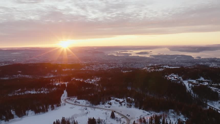 A mountainous landscape in winter at dusk featuring snow covered slopes and valleys dark evergreen forests and a winding road all under a sky with a bright sunburst effect at the horizon at sunset