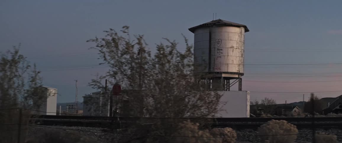 As trees and shrubs whiz by a water tower with a building beneath it is revealed next to rural train tracks at sunset from a moving vehicle
