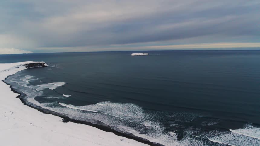 Waves foam as they approach a snow covered shore under an overcast sky on a winter day
