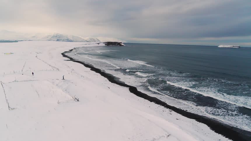 A snowy landscape stretches to a dark sandy beach where ocean waves roll in under a cloudy sky with a small figure standing alone on the beach