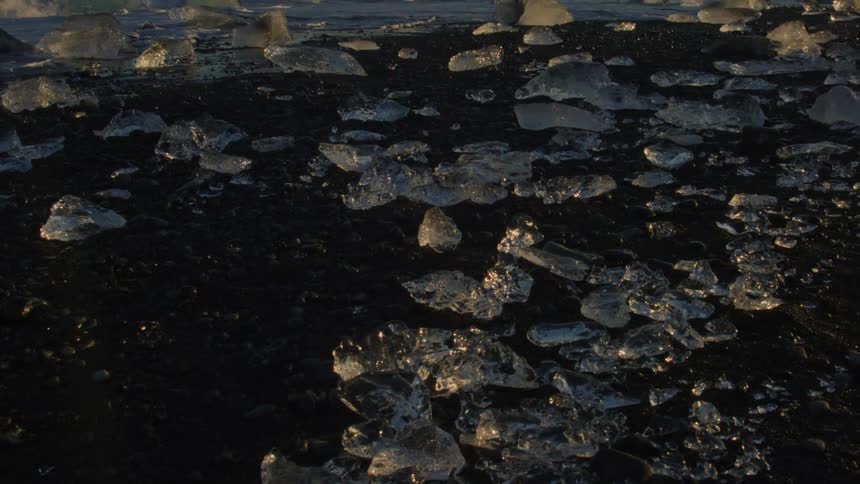 Fragments of transparent ice scattered over a dark sandy beach with waves in the background