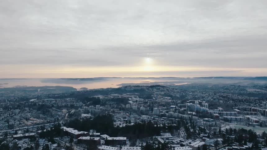 An aerial view of a cityscape with snowcovered buildings and trees surrounded by a vast landscape of hills and water bodies under a cloudy sky with a hint of sunlight breaking through
