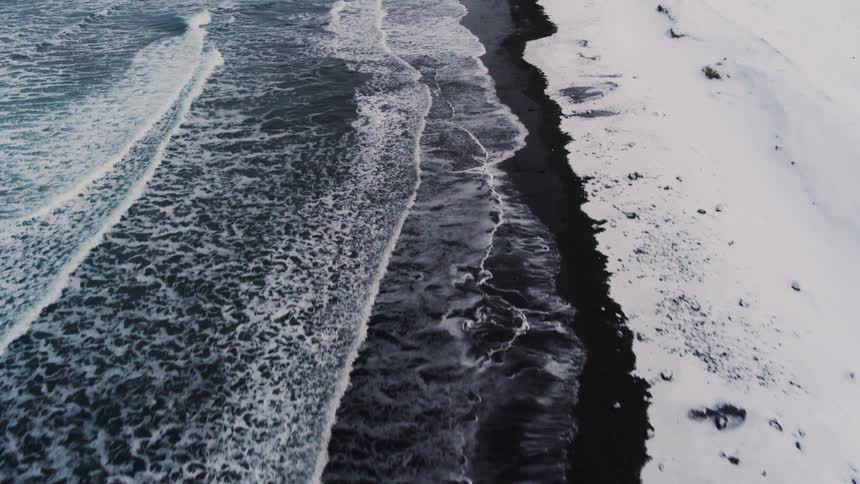 Aerial view of a black sand beach with foamy sea waves contrasting with the snow accumulated on the coast