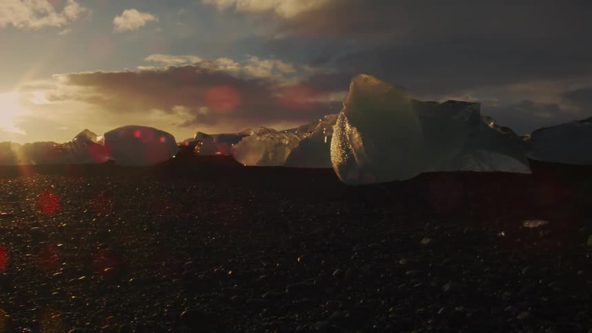 A scenic view of a rocky shoreline with large icebergs scattered across the ground illuminated by the setting sun