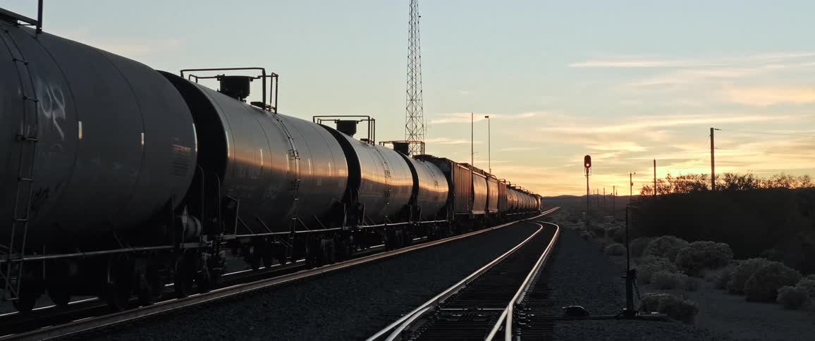 A line of dark train cars stretches into the distance along a track curving through a barren landscape at sunset with a tall radio tower in the background