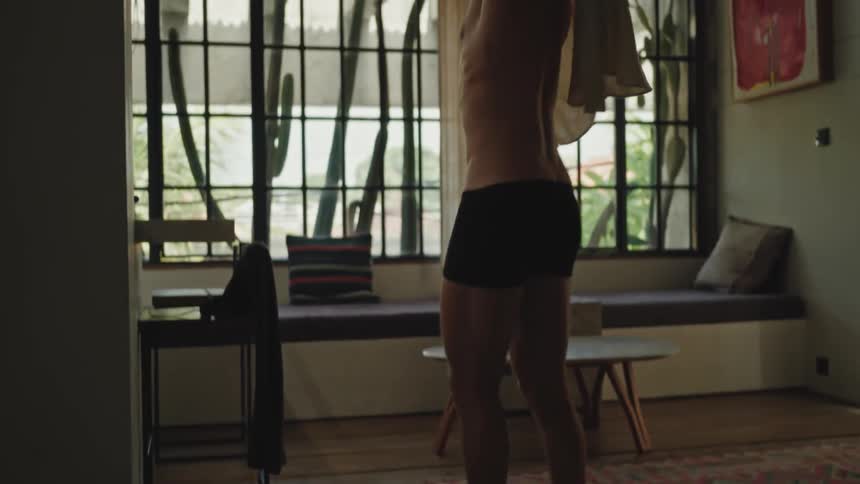 A shirtless man walking into a living room and putting on a tshirt while looking straight ahead next to a window