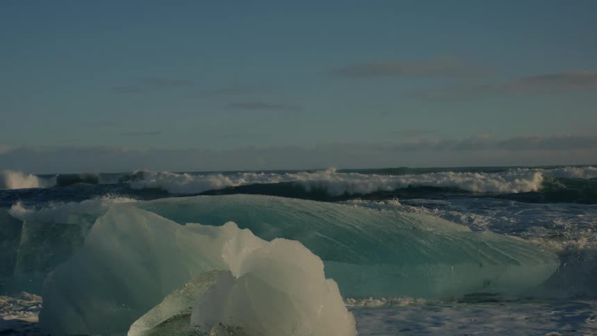 Ocean waves crash against blocks of crystal clear ice in an arctic landscape
