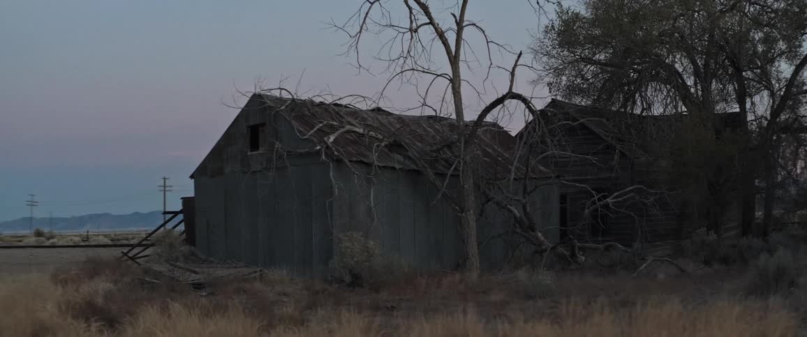 An old weathered barn with a dilapidated roof stands silhouetted against a fading sunset sky surrounded by dry overgrown fields