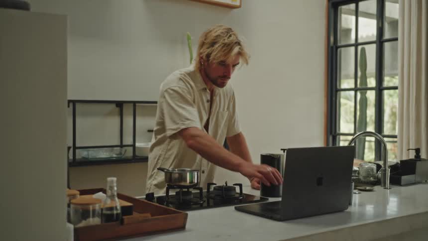 A man with blonde hair cooking at home while using a laptop in the kitchen