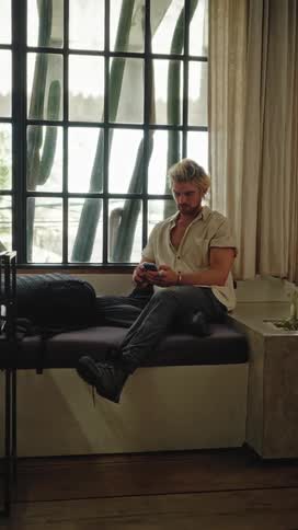 A man sits on a couch texting on his phone next to a window with cacti outside
