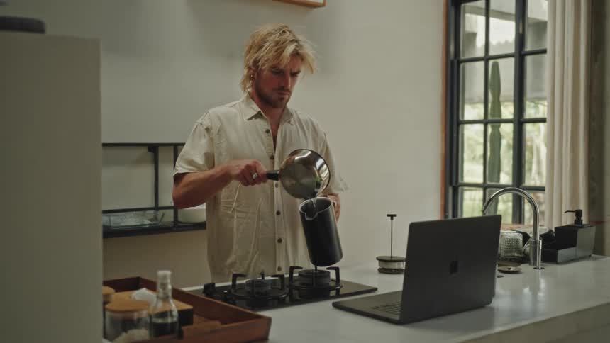 A blond man standing in a kitchen and is already passing water that is in a pan to a kettle and in front of him there is an open laptop