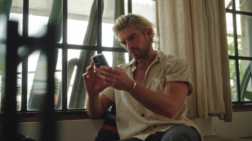 A blond man is sitting on a sofa there inside a window and he is fiddling with his cell phone