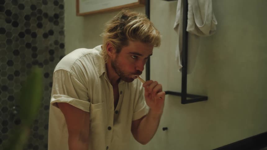 Blonde male brushing his teeth standing over bathroom sink