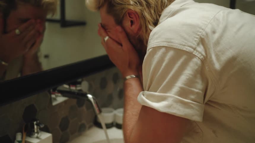 A blond man is using water from a sink tap to clean his face looking in the mirror