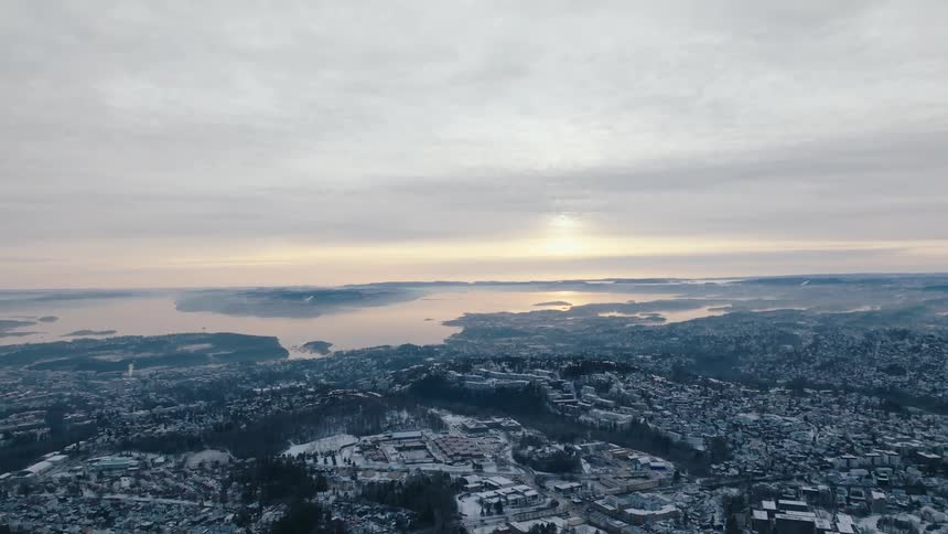A wide aerial view shows a snowcovered cityscape stretching to a distant horizon over a misty body of water