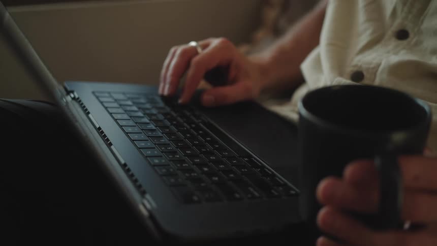 A person types on a black laptop while holding a black mug partially visible suggesting a casual or work from home setting