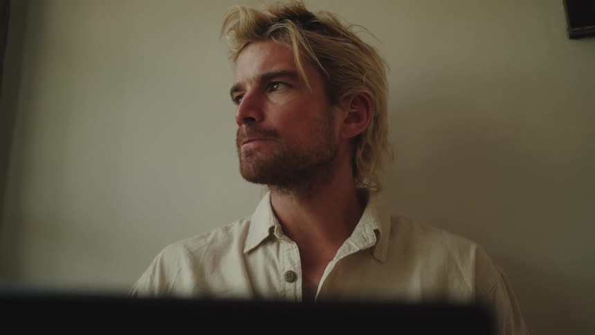 A blonde man in a white shirt drinks from his black cup while using a laptop