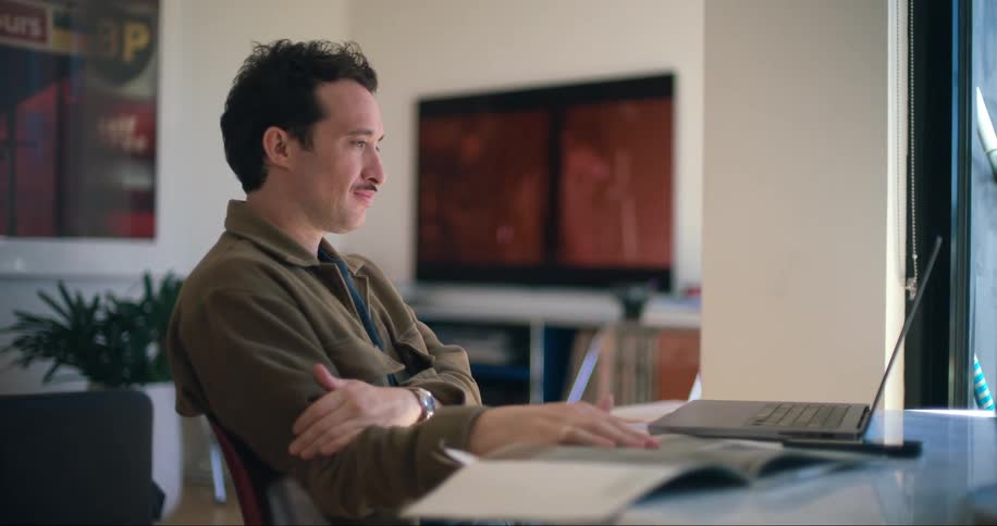 Man sitting turning the page of a book and facing a laptop on the table