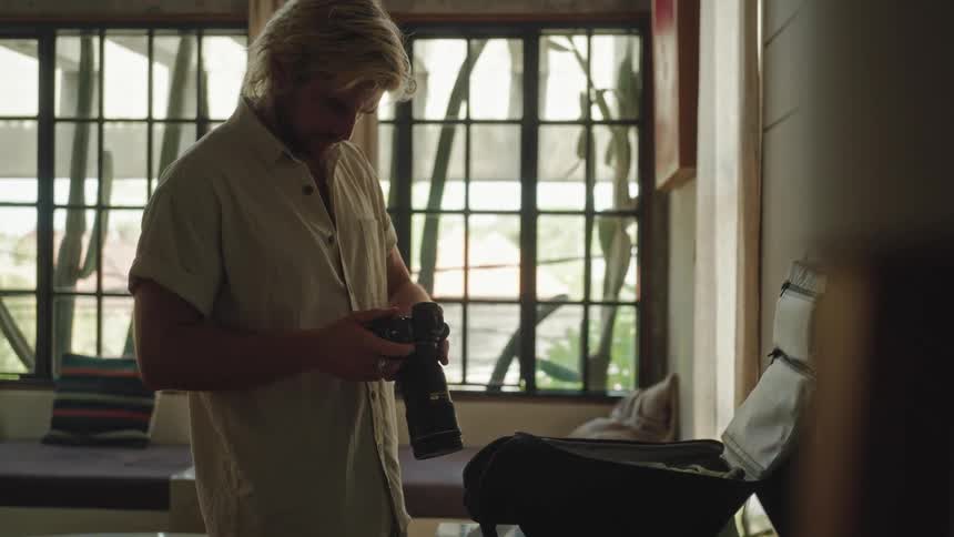 A blonde man wearing a light colored shirt stands indoors folding clothes before taking a camera and placing the items into a bag with a window and a chair visible in the background
