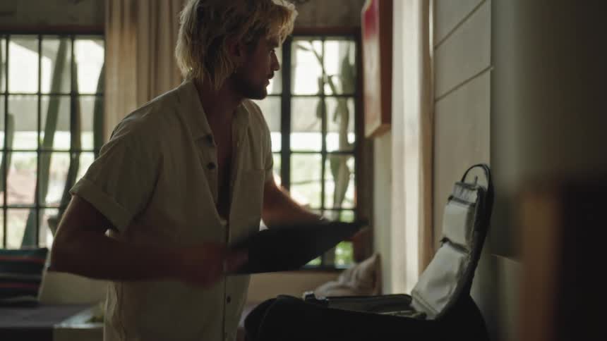 A blond man is standing in front of a table and putting his laptop away inside a backpack