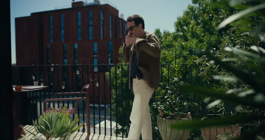 A man dressed casually in a brown jacket and light trousers is standing on a balcony talking on the phone with a modern building and greenery in the background