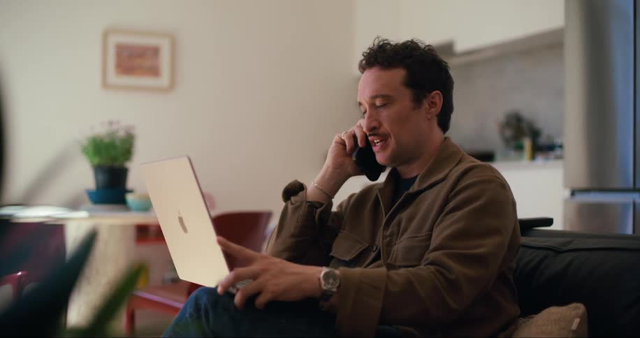 A man with a mustache wearing a brown jacket jeans and a wristwatch is sitting in a cozy living room smiling as he talks on the phone while holding a laptop