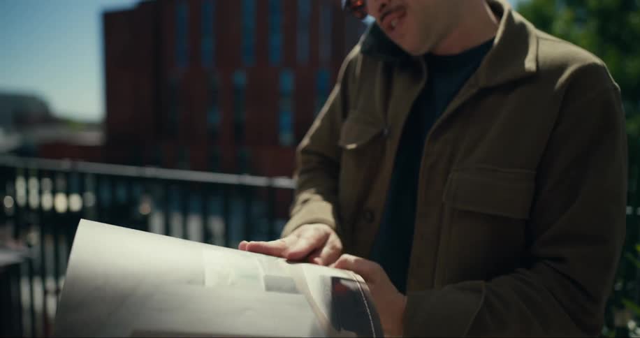 A man stands outdoors on a balcony while talking on his smartphone and looking at a magazine
