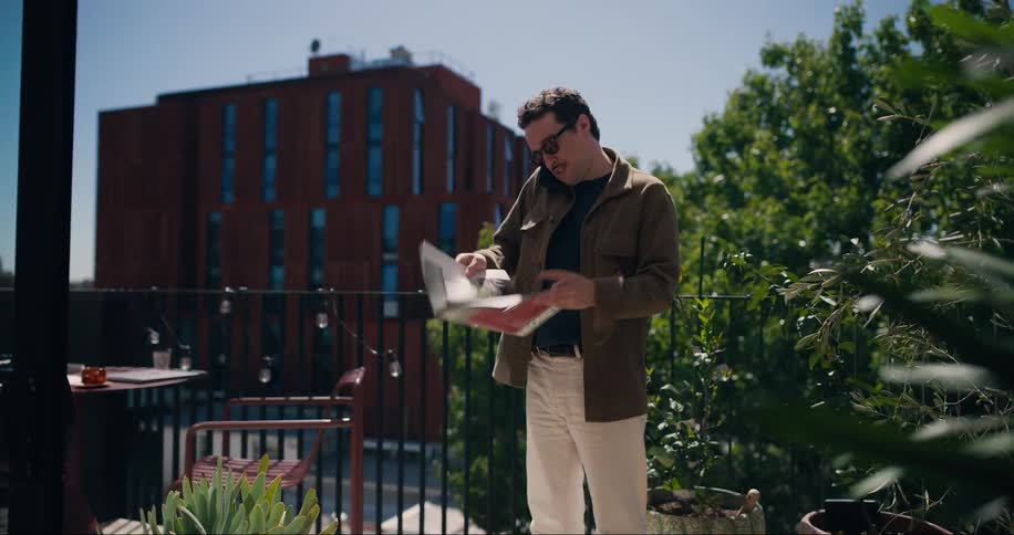Man walks around an outdoor cafe looking at a magazine while talking on the phone