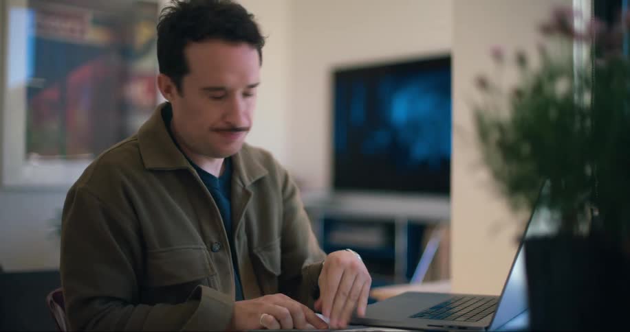 A man is failing a paper while he is reading it and is fiddling with his laptop which is on the table next to him